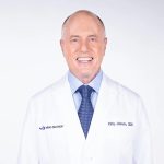 Smiling man in a white doctor's coat with a tie, representing a dental professional. Background is plain white.
