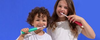 Two children with toothbrushes smile at the camera against a blue background, promoting good dental hygiene.