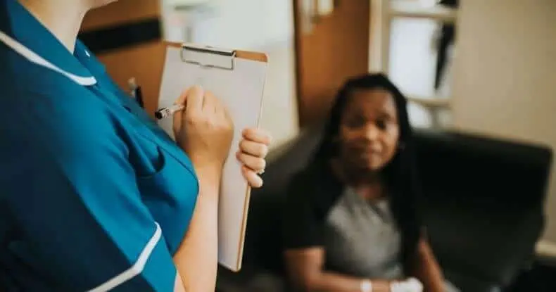 A nurse in scrubs holds a clipboard while speaking to a patient seated nearby in a medical setting.