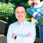 Man with a well-groomed appearance smiles confidently, arms crossed, with cartoon characters behind him amidst greenery.