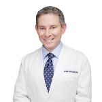 Smiling doctor in a white coat and blue tie, posing confidently against a plain background.