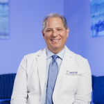 Smiling man in a white lab coat with a blue tie, sitting in a modern, softly lit office space.