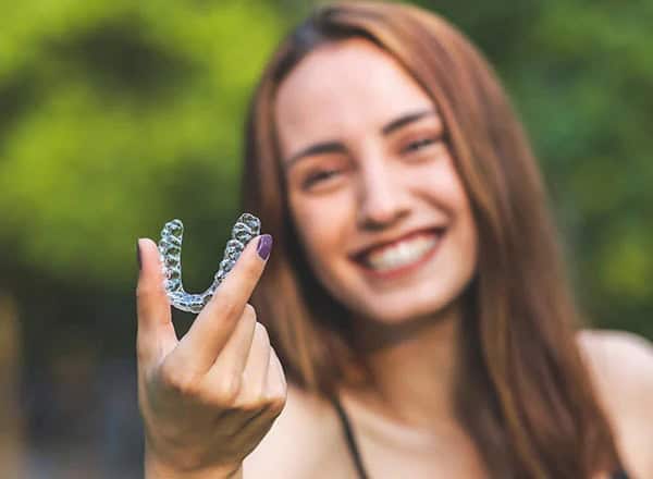 Smiling woman holding a clear orthodontic aligner against a blurred outdoor background.