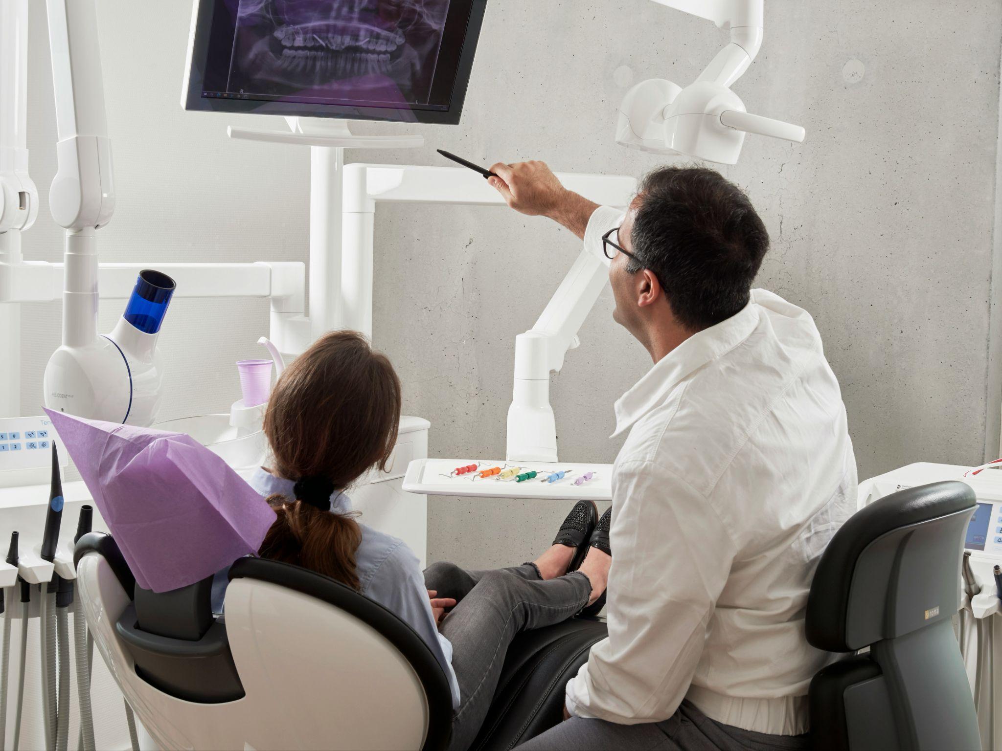Dentist pointing to a dental X-ray on a screen while explaining it to a patient in the exam chair