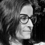 Close-up profile of a woman with long dark hair and glasses, set against a blurred background of greenery.