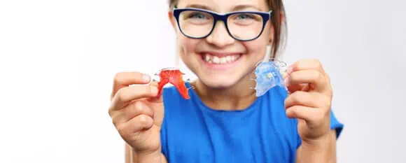 petite fille souriante et portant des lunettes tenant deux appareils dentaires de type faux-palais dans ses mains