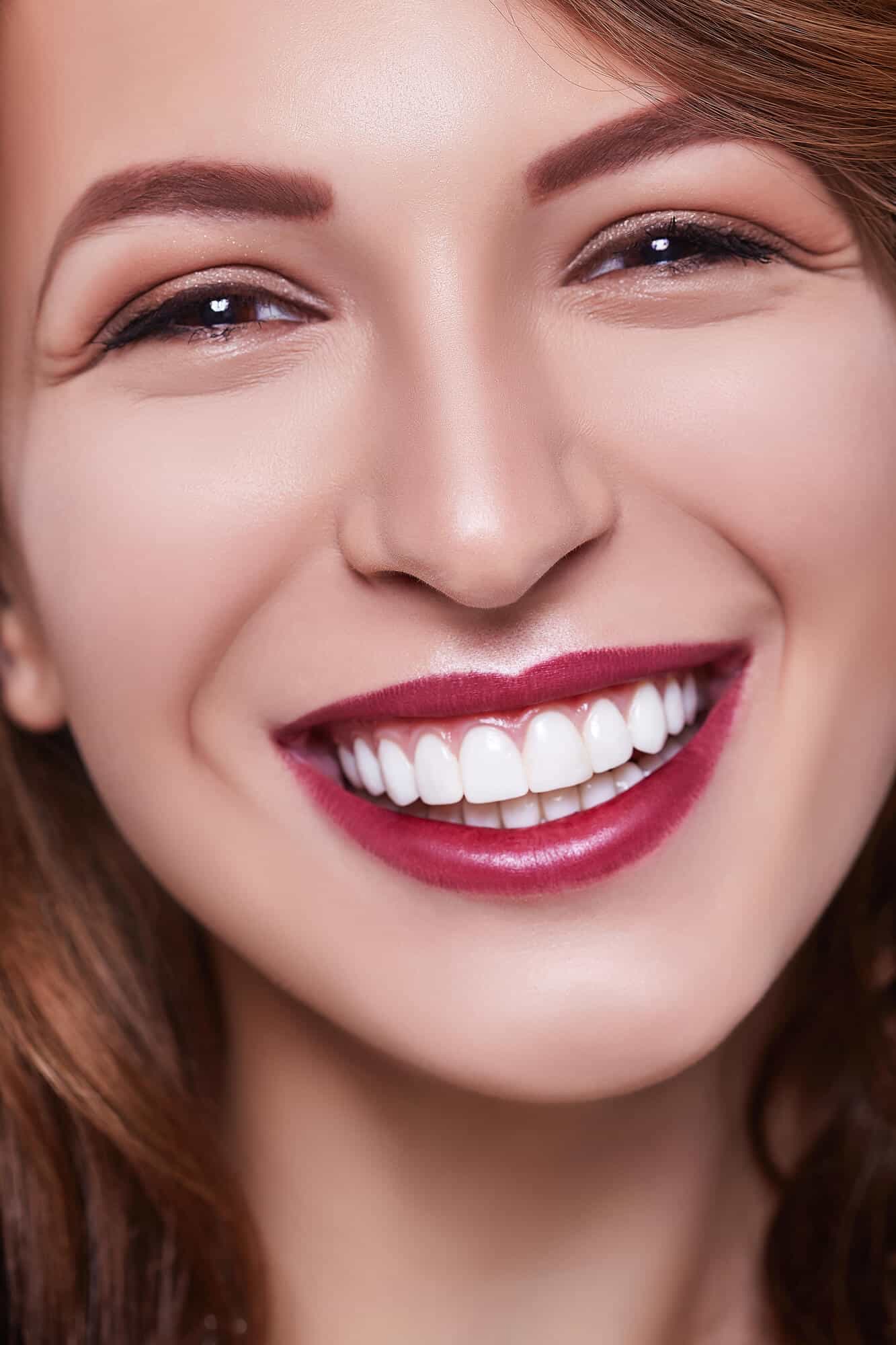 Close-up of a smiling woman with long hair, flawless skin, and deep red lipstick highlighting her bright, white teeth.
