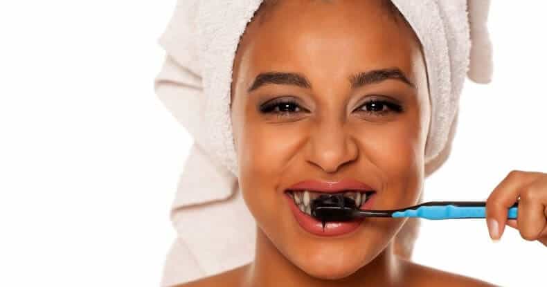 Smiling woman with a towel on her head brushes her teeth, showing a black toothpaste on her toothbrush.